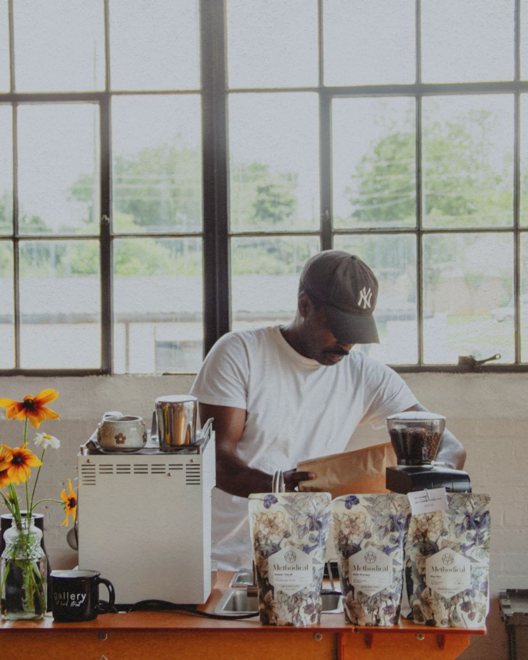 Person working behind a counter with coffee equipment and bags in a bright room with large windows.
